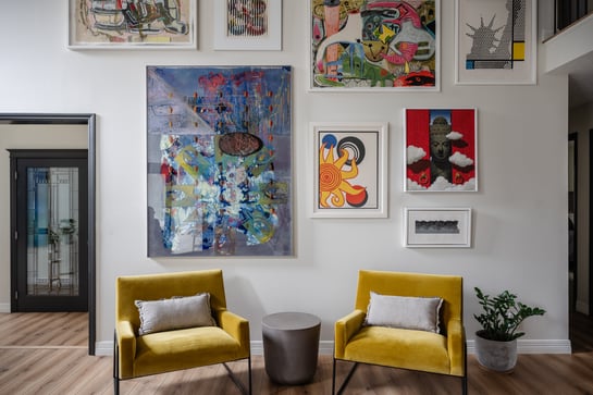 Interior living space with artwork hung on white walls and 2 yellow chairs with a side table in between.