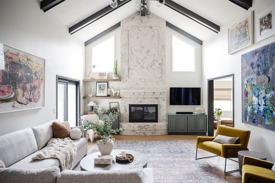 Living room with large fireplace in the center wall of the room. The walls and fireplace are white with a grey couch and colorful artwork on walls.