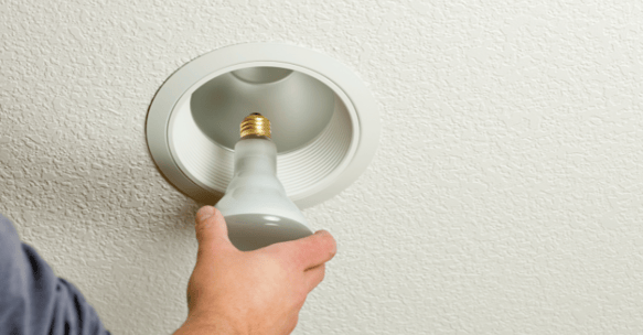 Man removing light bulb from recessed light fixture on white ceiling.
