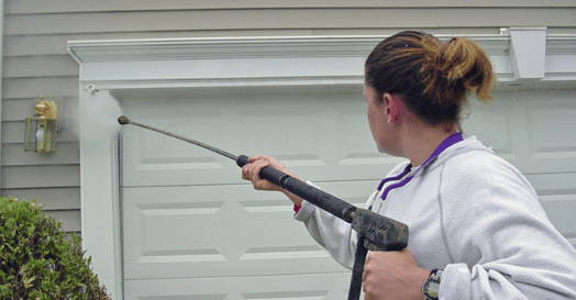 Woman power washing exterior of home.