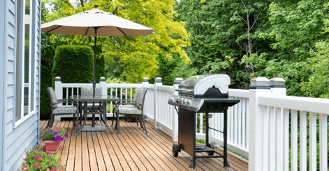 Deck with white railings and warm brown stain on wood. Deck has patio furniture and grill on with flowers along the exterior of the house.