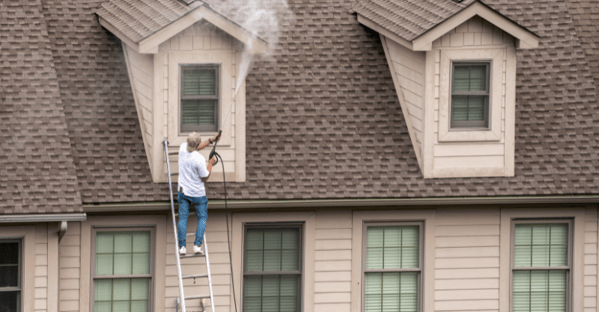 Man power washing upper level of exterior of home on ladder.