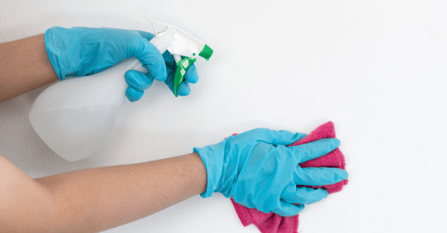 Person cleaning white wall paint with a clear liquid and a pink microfiber cloth, wearing gloves.