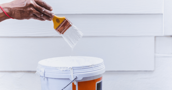 Person pulling paint brush out of paint bucket filled with white exterior house paint.