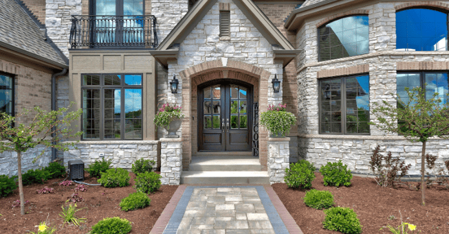Exterior entrance of large stone home with big windows and landscaping lining the walkway.