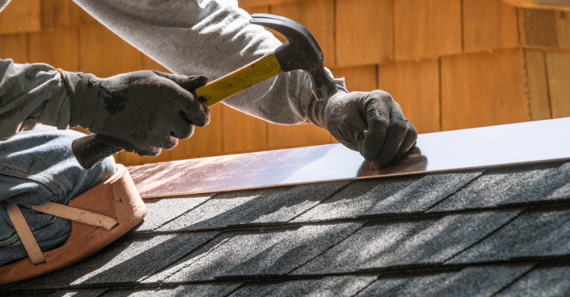 Person hammering a nail into roof shingles on the top of a house in Omaha.