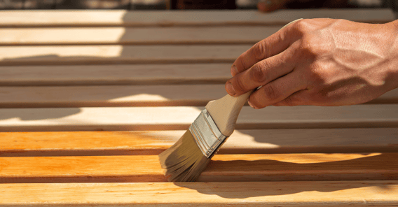 Person brushing warm brown deck stain on to thin wood planks on a deck.