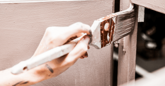 Person painting cabinet boxes and doors with a brush and light brown paint.