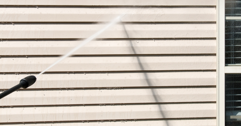 Power washer hitting the side of a house with vinyl siding painted a light beige color.