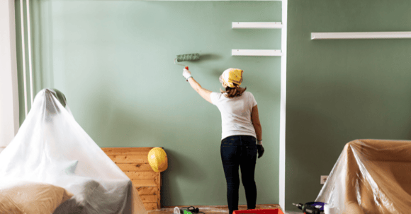 Woman painted wall sage green with painting supplies around her.