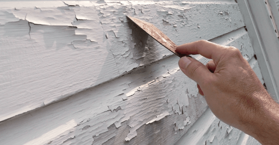 White exterior paint on house that is peeling, someones hand is using a scraper to scrape the peeling paint off.