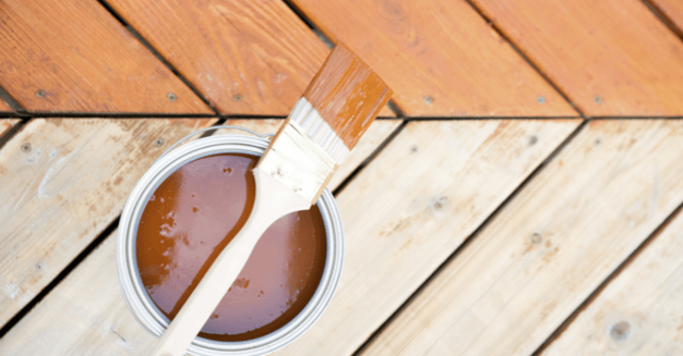 Half of wooden deck stained light warm brown and half unstained with a quart of stain and a brush sitting on top.