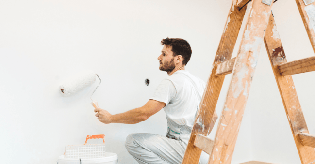 Painter rolling white paint onto wall in a room.