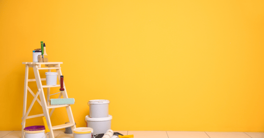 Wall painted bright yellow with paint gallons and supplies on the floor and a ladder next to the wall.