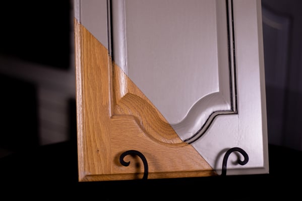 Cabinet door painted off white, beige, with grain lightly showing through the paint. Corner of the door shows the untouched natural wood before painting.