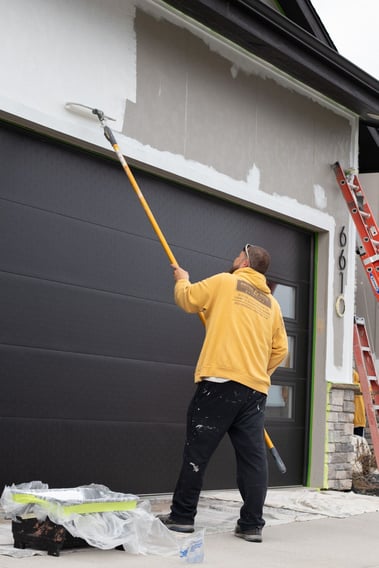 Painter rolling white paint on siding with white paint above garage in Omaha, NE.