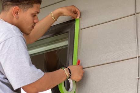 Painter taping off window trim on exterior siding to prepare to paint.