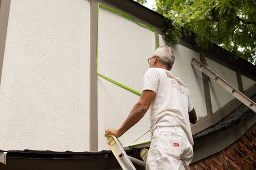 Painter peeling green painters tape from panels of stucco on house.