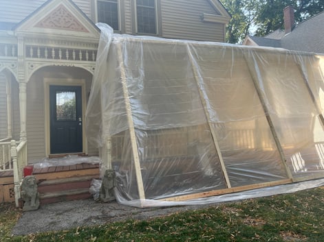 Exterior of home that has lead paint being peeled off to be repainted a new color. There is a plastic wall with wooden beams against the home.