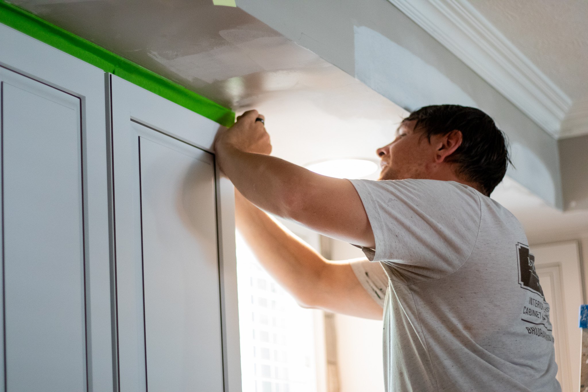 Painter applying green frog tape to upper cabinets in a kitchen that were painted white. 