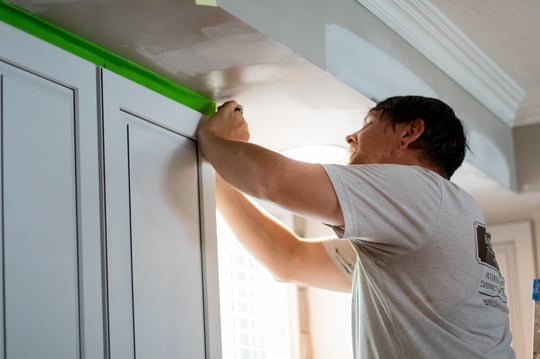 Brush & Roll Painting employee taping off edges of white kitchen cabinets before re-painting the walls around them.