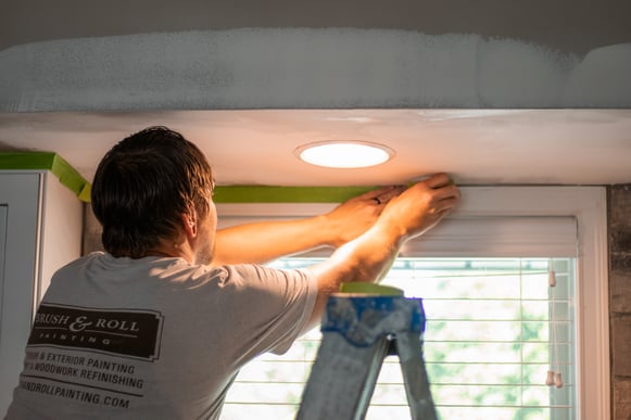 Painter applying tape along a window frame before painting a ceiling in a kitchen.