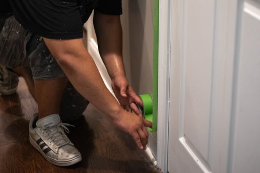 Painter rolling green tape onto white baseboards before painting the wall inside a home in Omaha, NE.