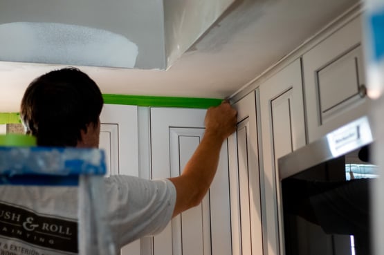 Painter applying tape on the upper cabinets painted white, along the top of the box frame in a kitchen..