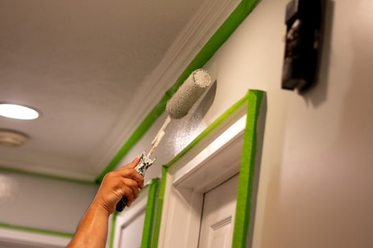 Painter rolling white paint onto a wall with green painters tape around edges.