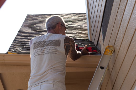 Brush & Roll Painting painter replacing siding on a beige colored house with dark roof shingles.