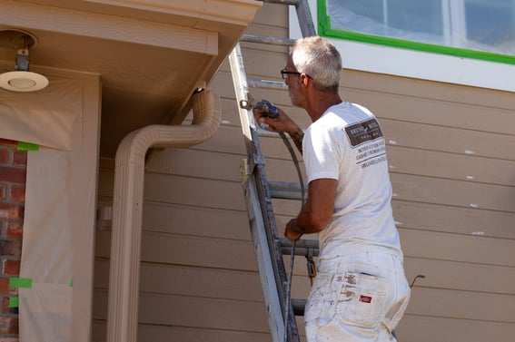 Painter spraying light brown paint against the outside of a house in Omaha, NE using Aura by Benjamin Moore.