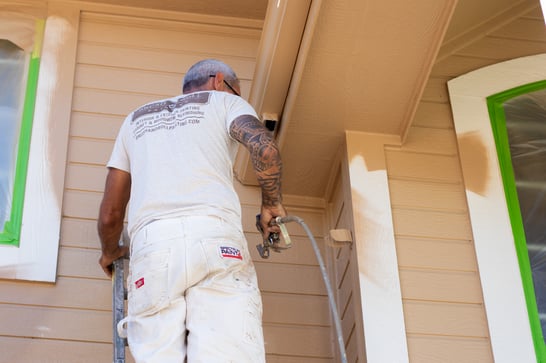 Painter spraying beige paint onto siding and fascia on a Omaha, home.