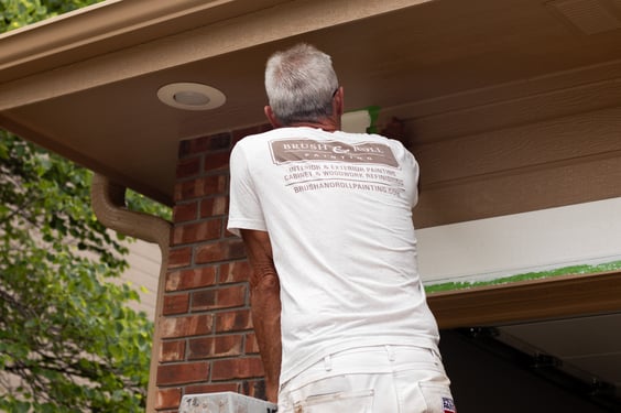 Exterior home painter brushing and repairing paint on the front side of a house above the garage.