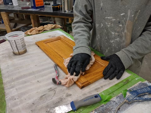 Cabinet painter cleaning a golden oak cabinet door with denatured alcohol and a rag.