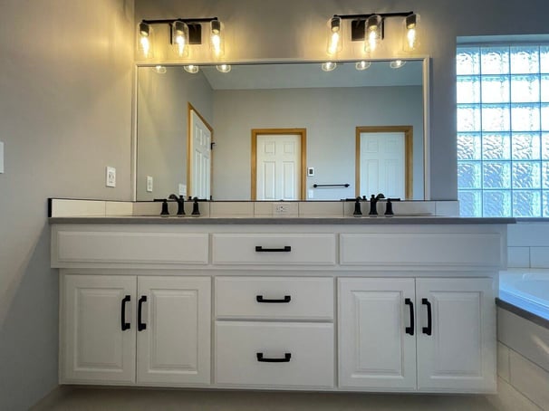 Bathroom vanity with 2 sinks with cabinets painted white with black handles.