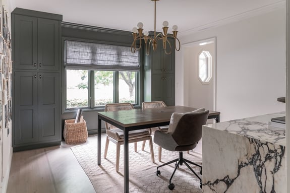 Interior home office with a desk in the middle of the room and sage green painted bookshelf along the back wall with a window.