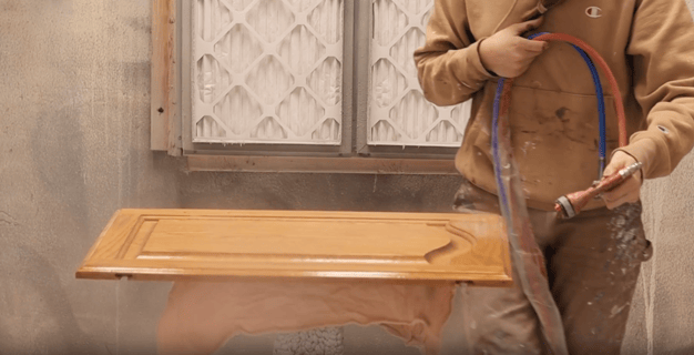 Painter spraying oak cabinet door with a clear resurfacer layer,