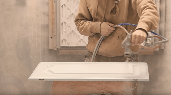 Painter spraying an oak cabinet door with white 2k polyurethane paint inside a spray booth.