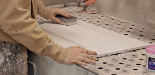Painter using a sanding block on a cabinet door with a coat of primer on it in the shop at Brush & Roll Painting.