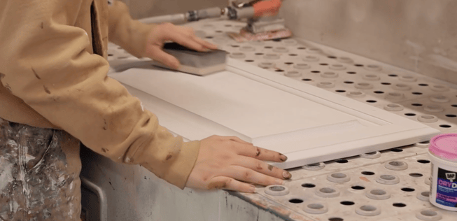 Painter using a sanding block on a cabinet door after spraying a layer of primer.
