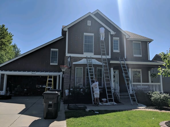 Painters on ladders against a house painted brown while doing prep work for exterior house painting.
