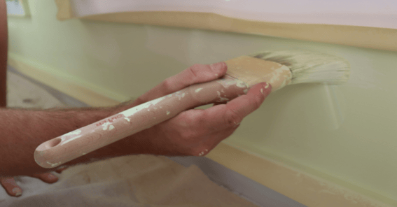 Person brushing light green paint on wall with tape along edges.