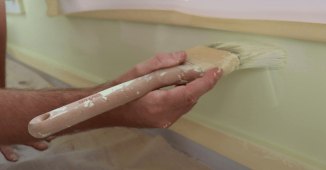 Person brushing green paint onto a wall under a window sill with painters tape on the trim.