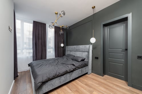 Grey-green painted accent wall with bed in middle of room. Window with floor length dark purple curtains and warm wood floor.