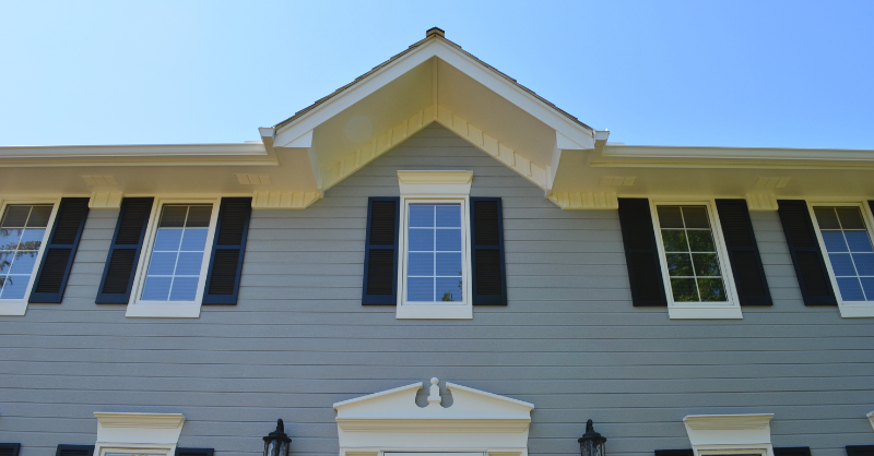 Upper view of the front of exterior home with siding painted blue, dark black window shutters and white window trim.
