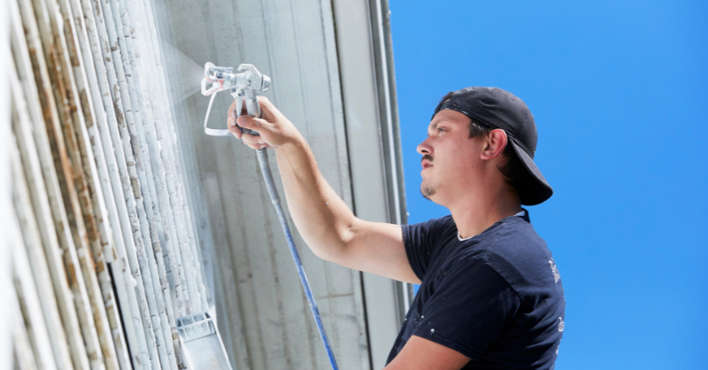 Homeowner DIY painting the exterior of their house white with a paint sprayer.