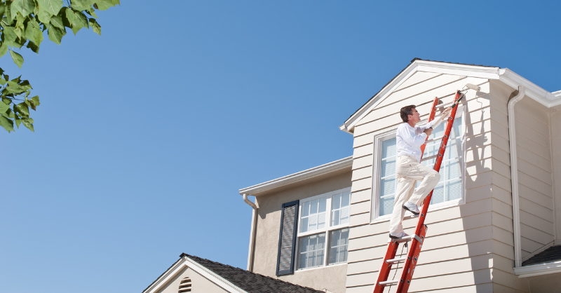 Exterior painter painting top arch of house beige with white trim.