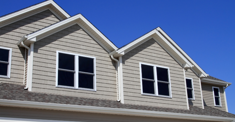 Top peaks of exterior of house with roof shingles and siding painted beige with white trim around roof and windows.