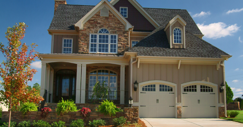 Exterior of home painted a light brown color with stone and white garage doors. Front yard has bushes and landscaping.