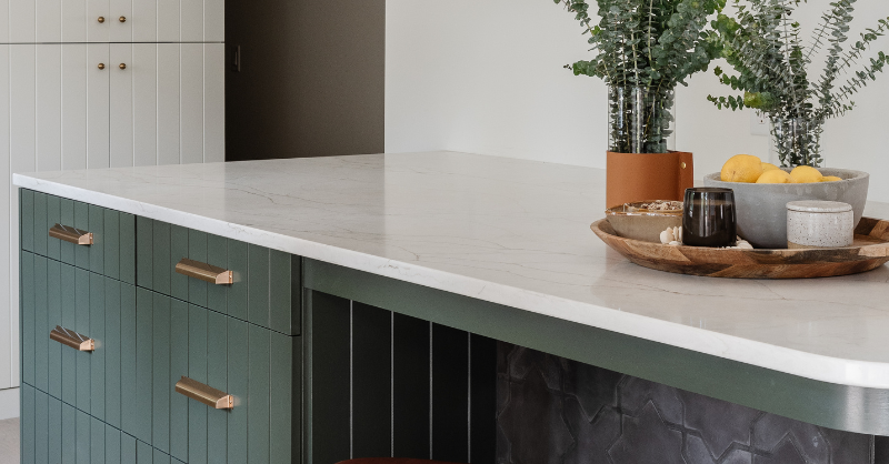 Kitchen with sage green island and gold handles on drawers with eucalyptus on countertop.